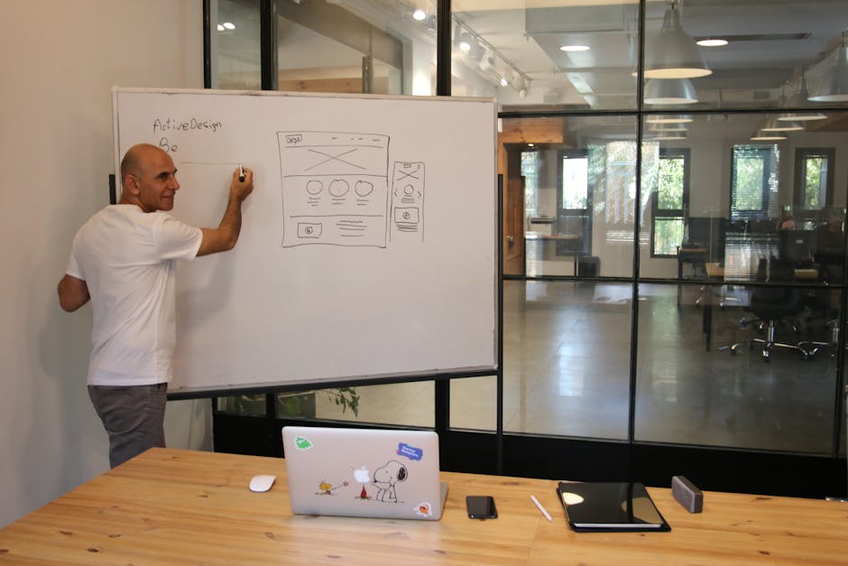 Side view of middle aged male leader in white t shirt writing on whiteboard during presentation in modern office space with glass walls