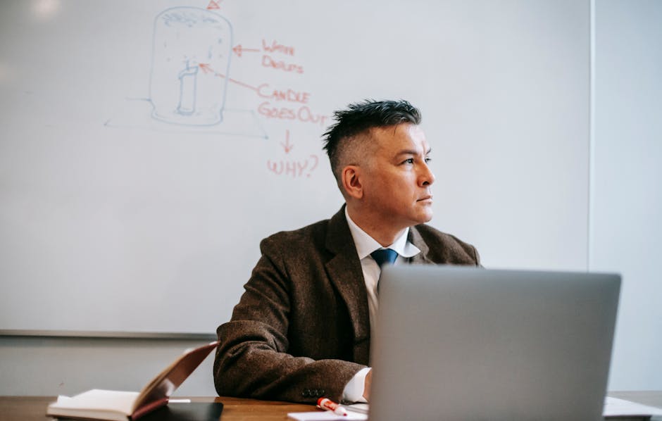 An adult man in formal attire working at a desk with a laptop, appearing thoughtful.